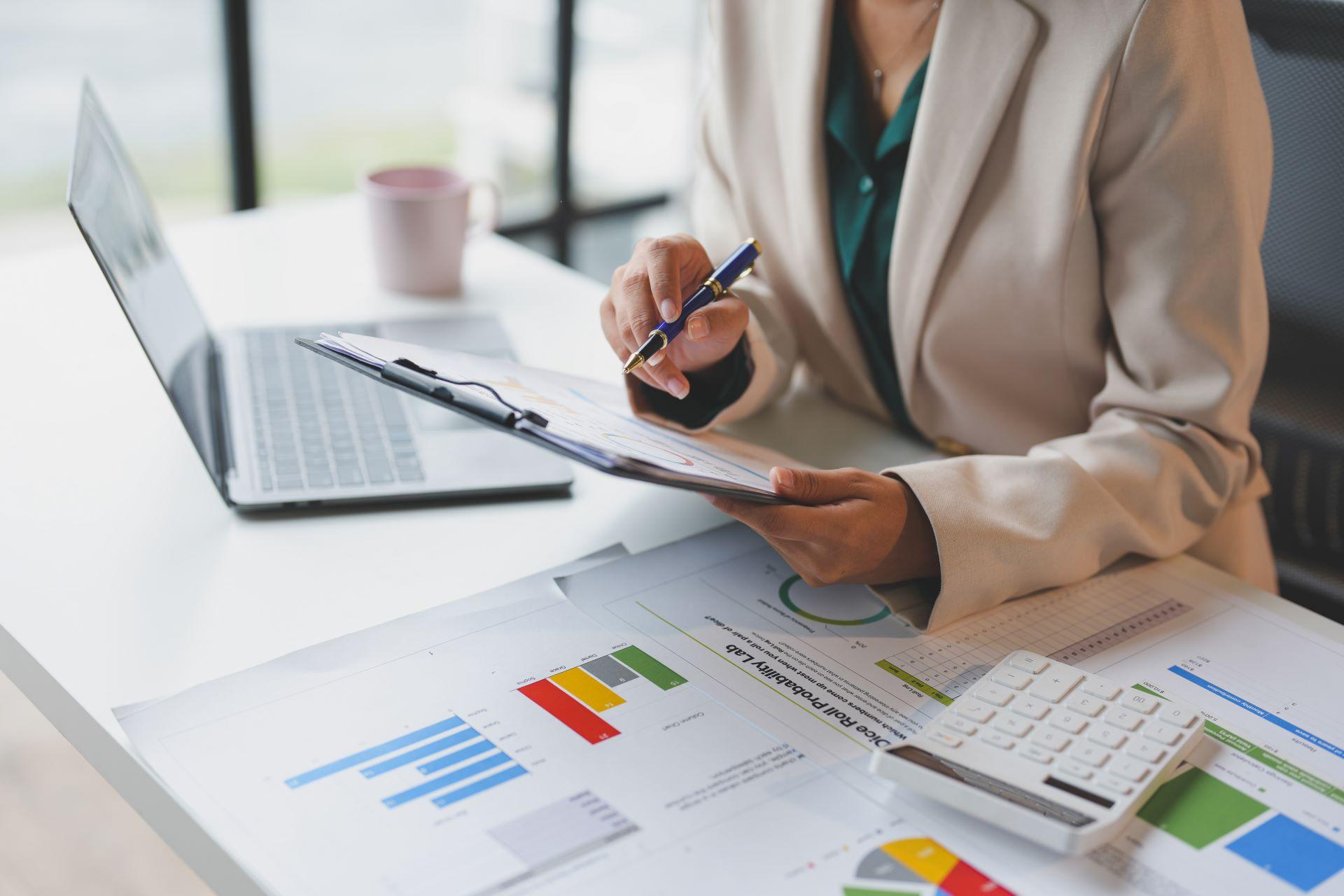 Eine Person in einem Business-Outfit überprüft Finanzunterlagen und Diagramme an einem Schreibtisch, unterstützt von einem Laptop und einem Taschenrechner. Das Bild symbolisiert die professionelle Steuerberatung durch einen Steuerberater in Neu-Ulm zur Optimierung der Finanzen.